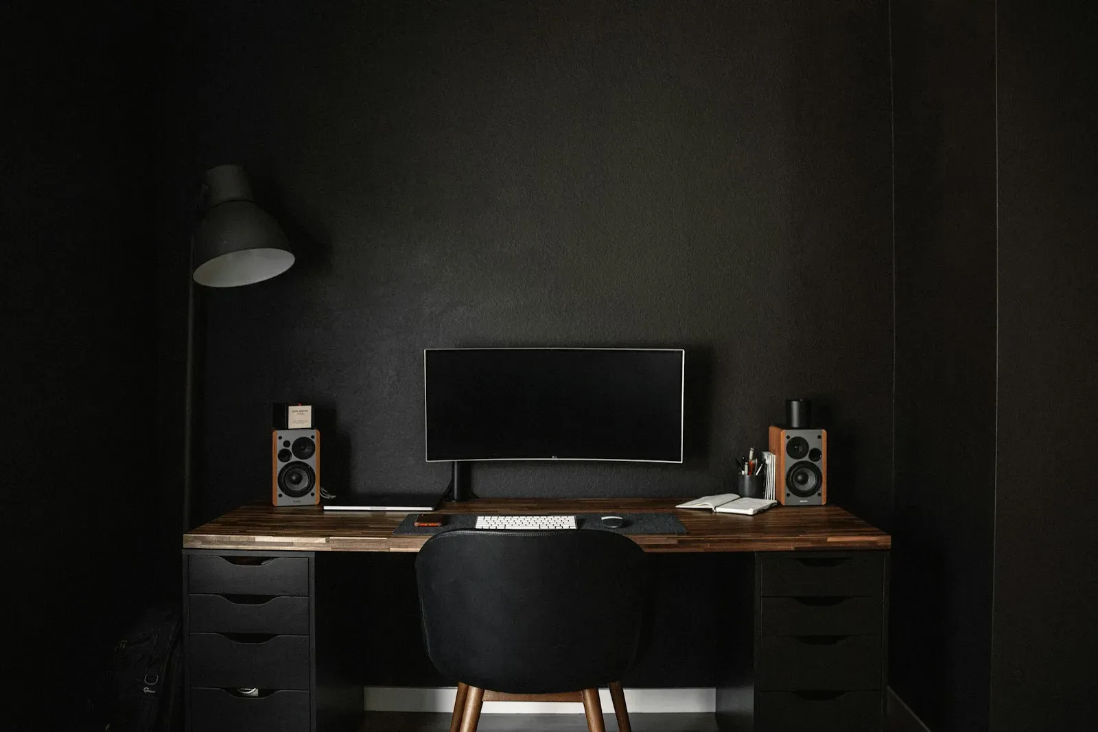 A dark, uncluttered workspace with a widescreen monitor, speakers and a keyboard